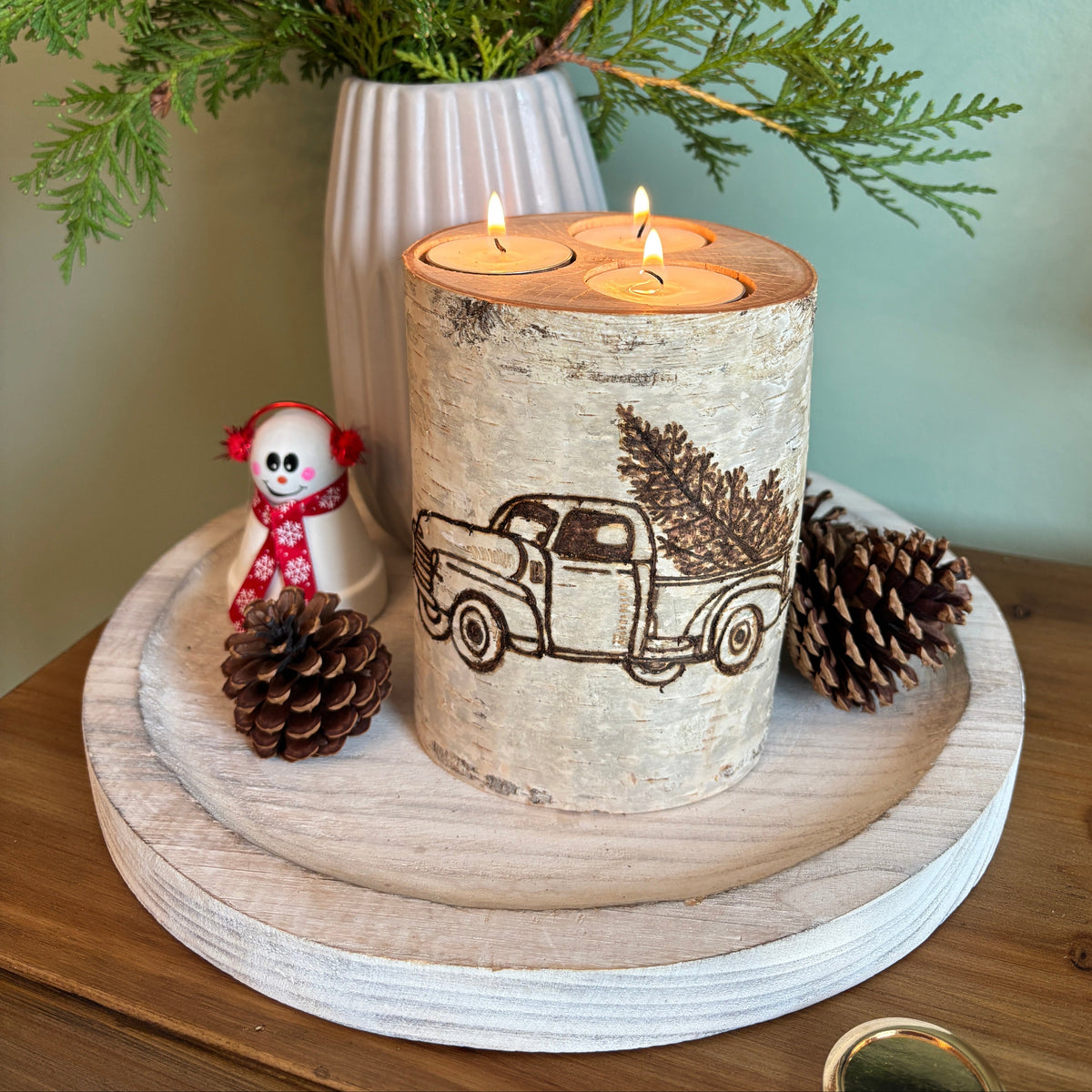 A rustic birch log candle holder with a printed truck design, holding three lit tealights, styled on a round wooden tray with pinecones, a small snowman figurine, and a white vase of greenery against a pale green wall.