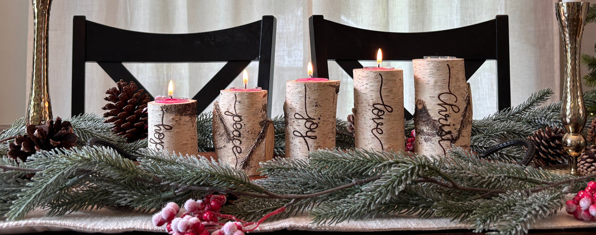 A festive holiday table centerpiece with five lit taper candles wrapped in natural birch bark, surrounded by pine branches, pinecones, and pink berries on a dark wooden table. Two tall white taper candles in gold holders stand on either side, with black chairs and sheer curtains in the background.