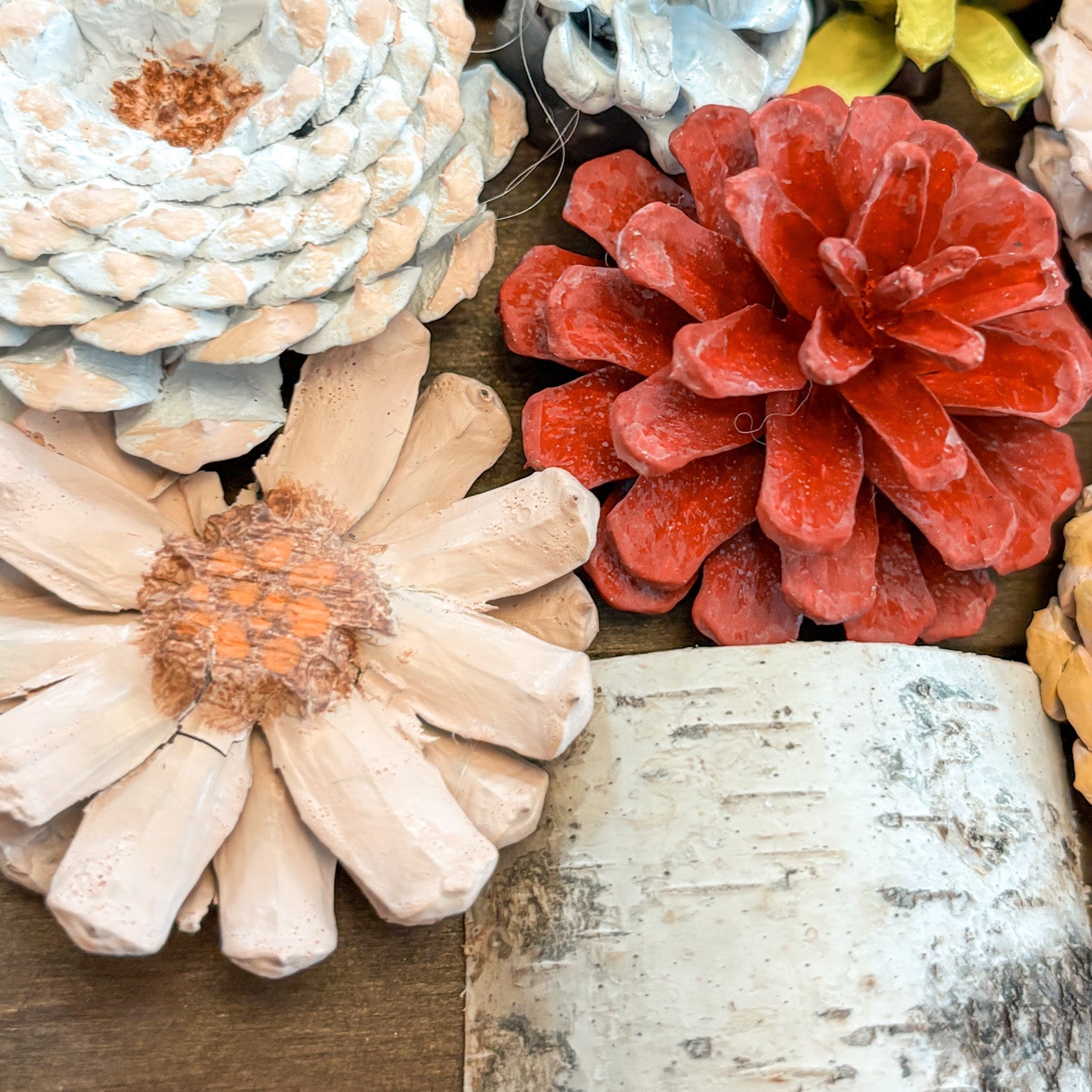 A close-up arrangement of decorative faux pinecones and flower-like pinecone ornaments in muted autumn colors, including pale blue, coral red, cream, tan, and yellow-green, with a birch bark texture at the bottom.
