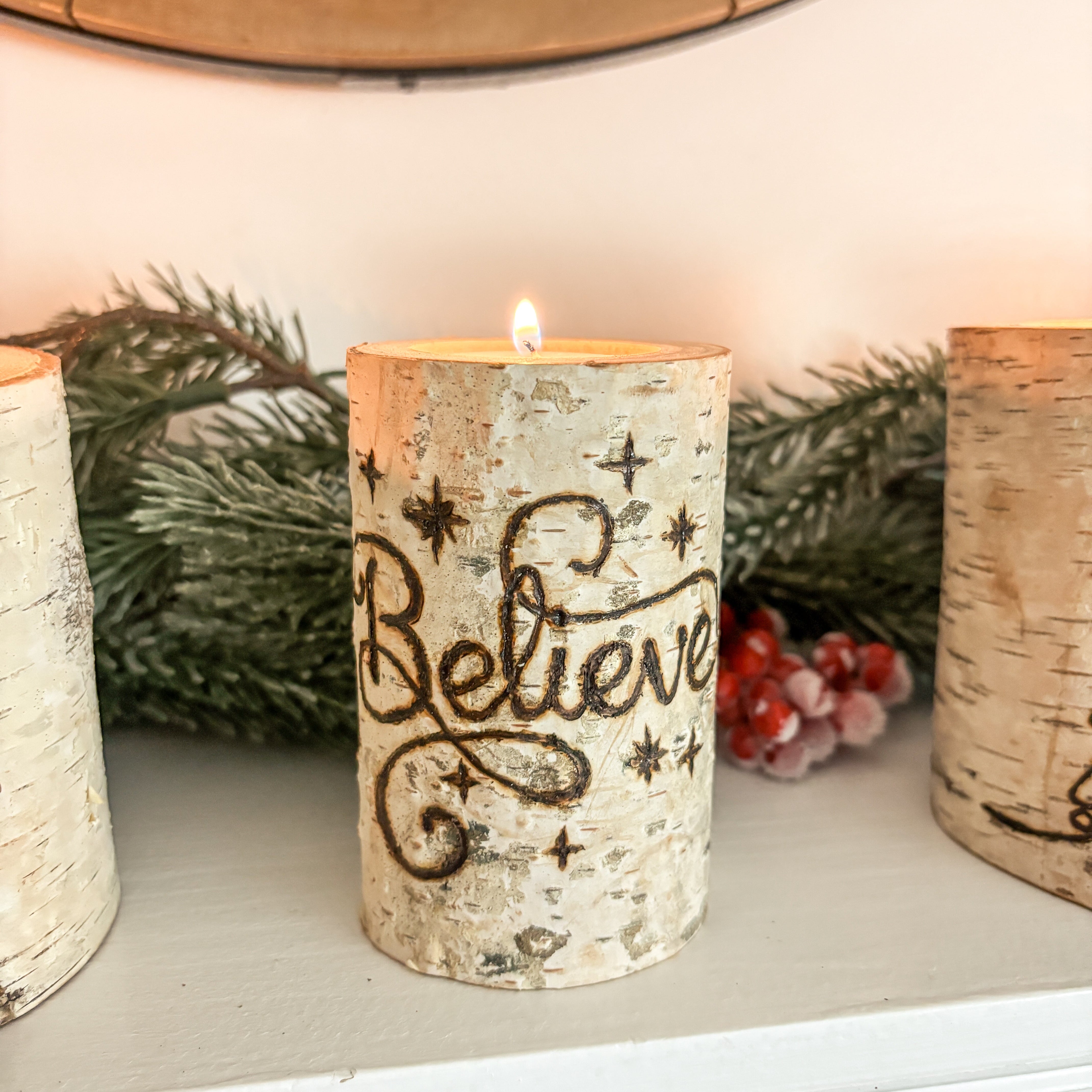 Festive candle with 'Believe' text on a decorative surface with greenery and berries.
