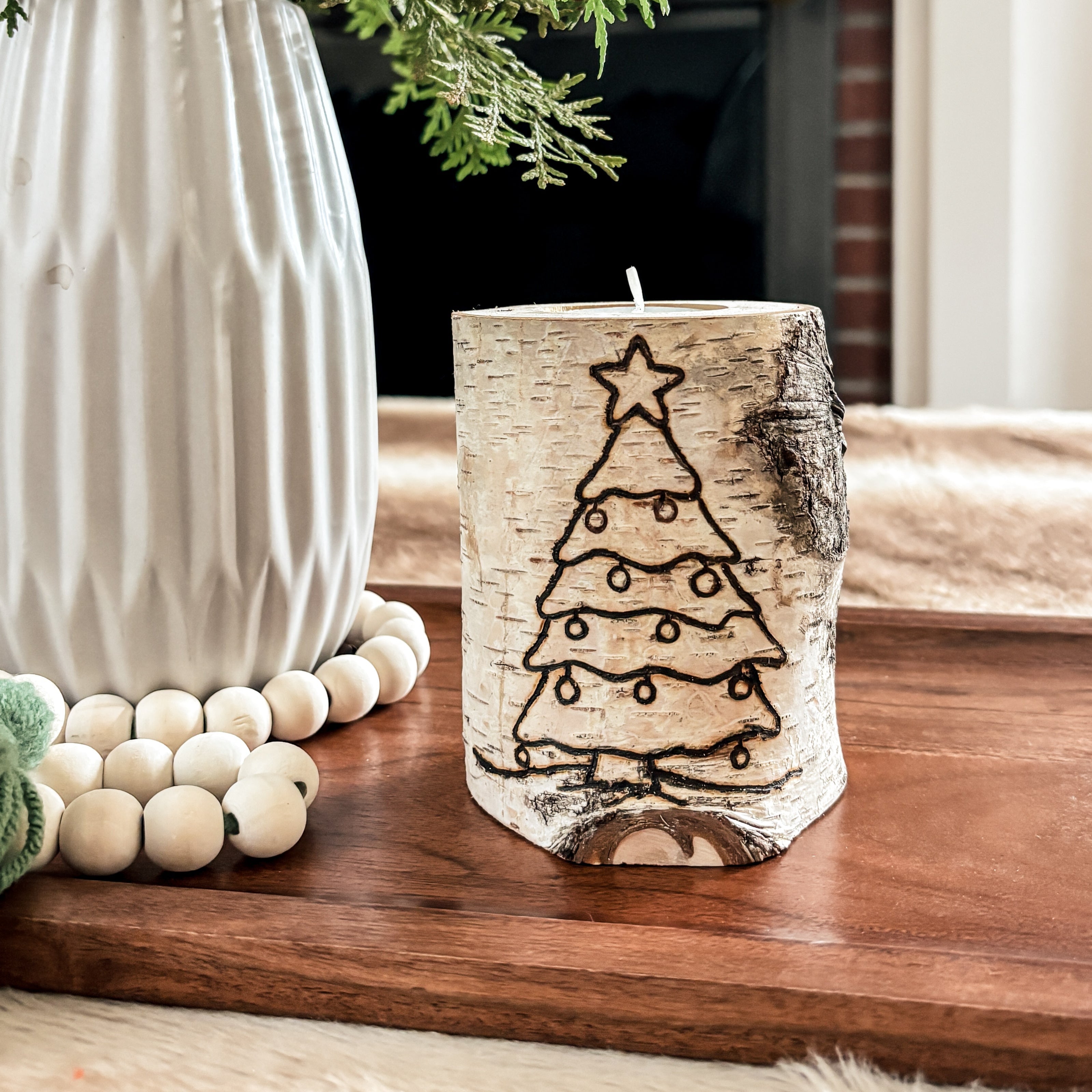 Decorative candle with Christmas tree design on a wooden surface, next to a vase with greenery.