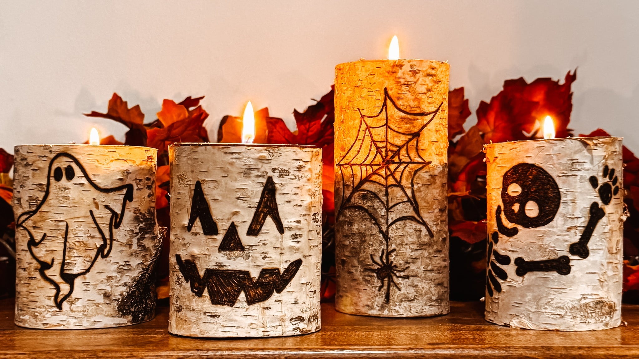 Four lit birch wood pillar candles with Halloween designs including a ghost, jack-o'-lantern, spiderweb, and skull, displayed on a wooden surface with autumn leaves in the background.