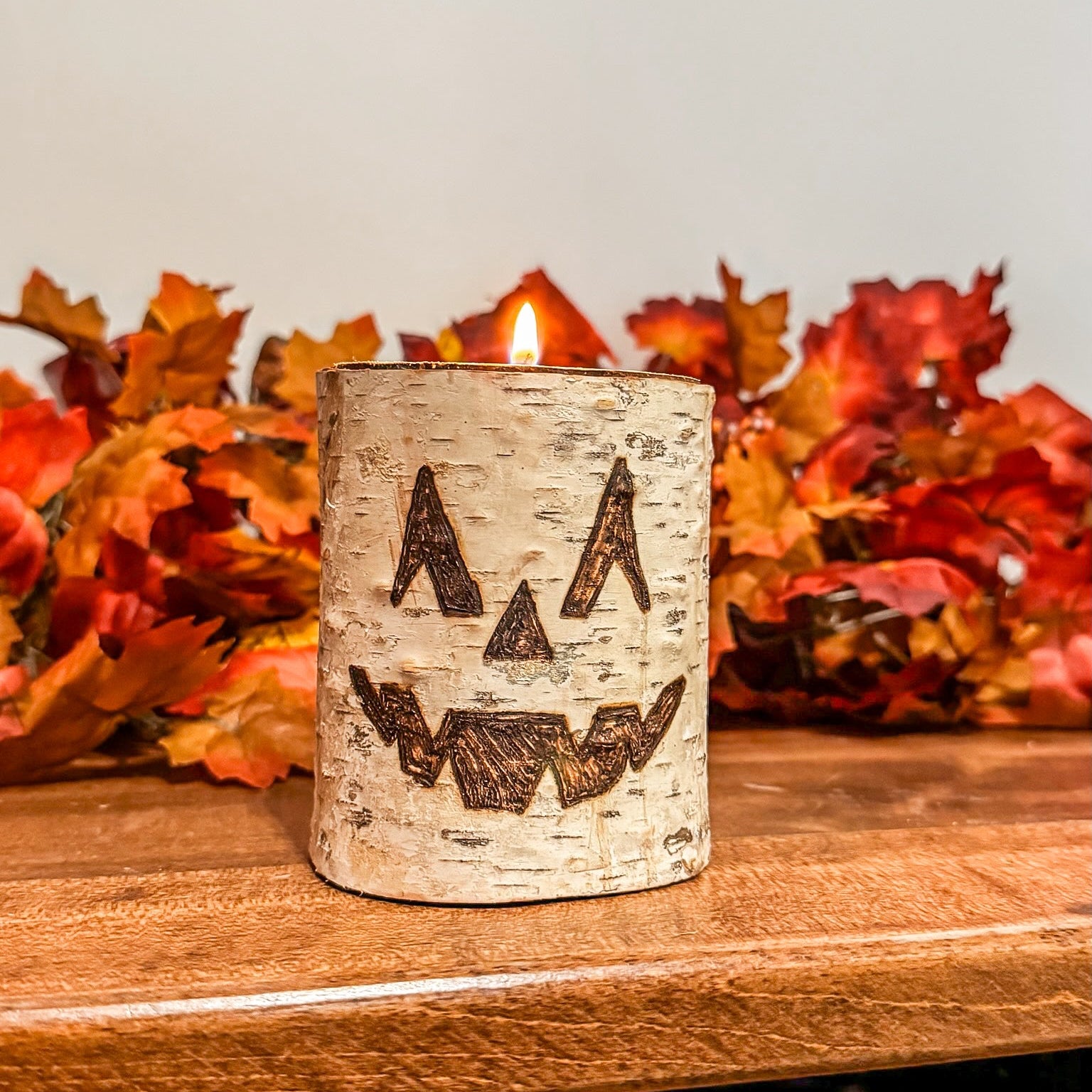 Birch bark candle with jack-o'-lantern design on a wooden surface with autumn leaves in the background.