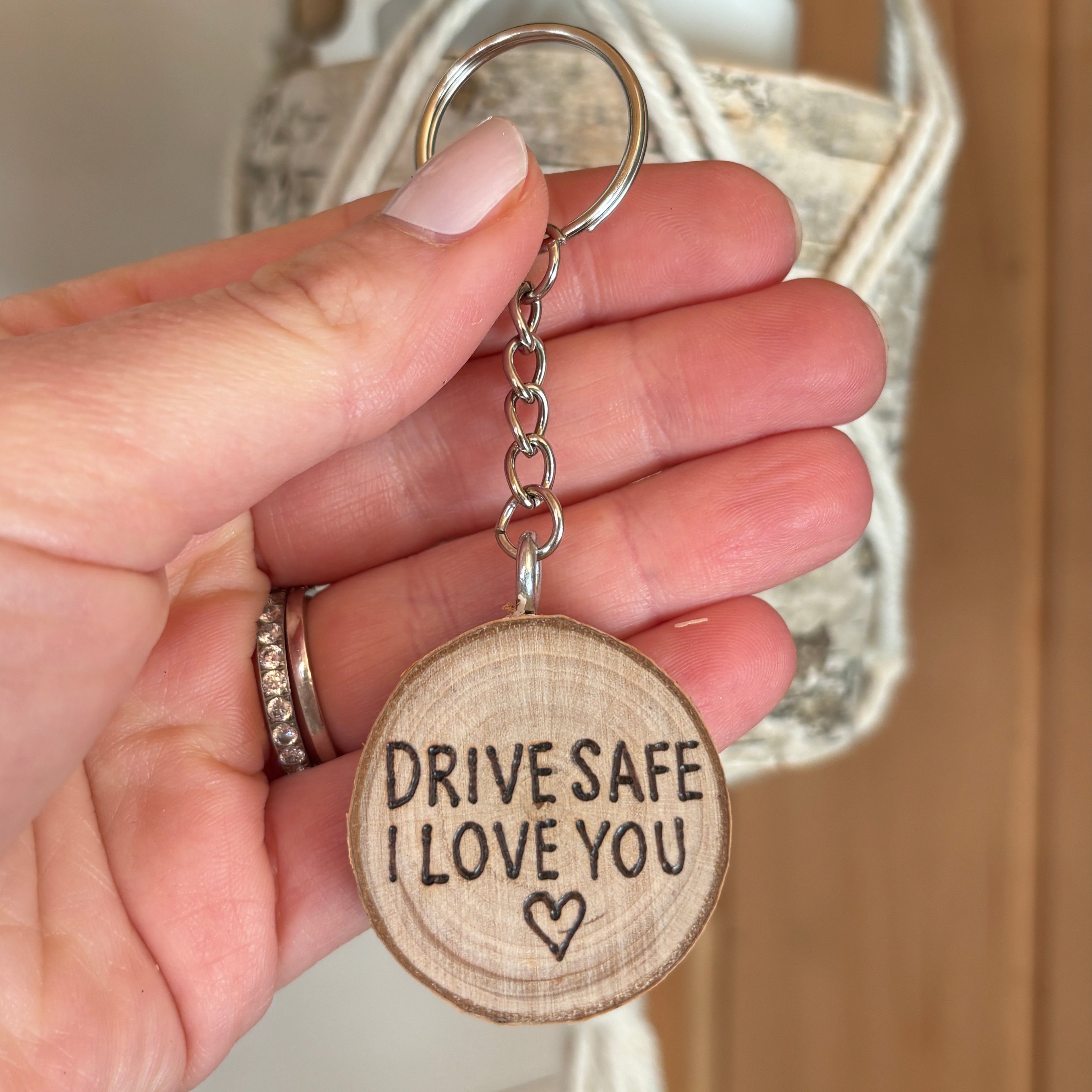 Hand holding a wooden keychain with 'Drive Safe I Love You' text and heart symbol, blurred background