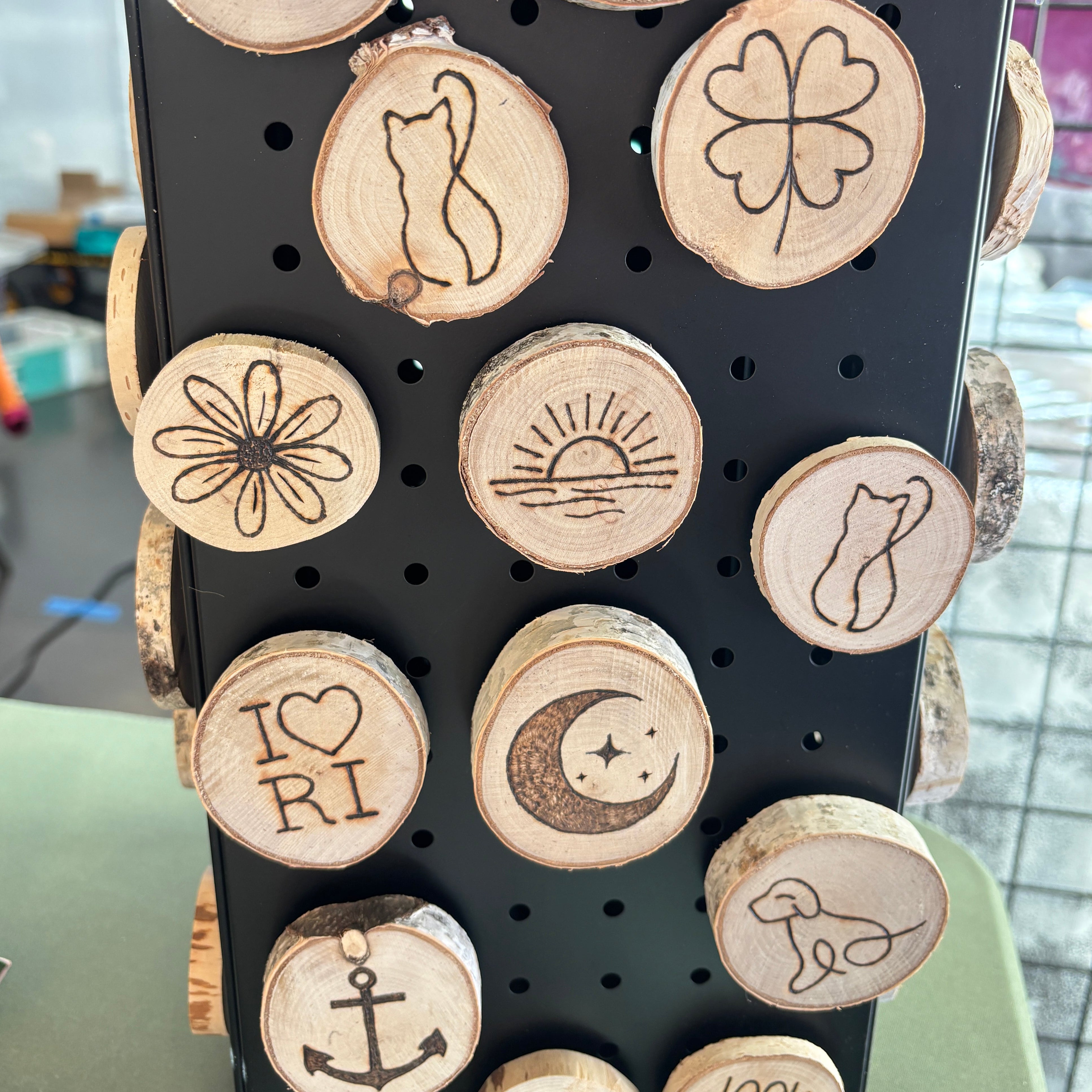 A display of round wooden magnets on a black pegboard, featuring various laser-engraved designs including 'I ❤️ RI', 'happy', 'LOOK WHAT I Made', a cat silhouette, a four-leaf clover, a flower, a sun, a crescent moon with stars, an anchor, and a dog silhouette.