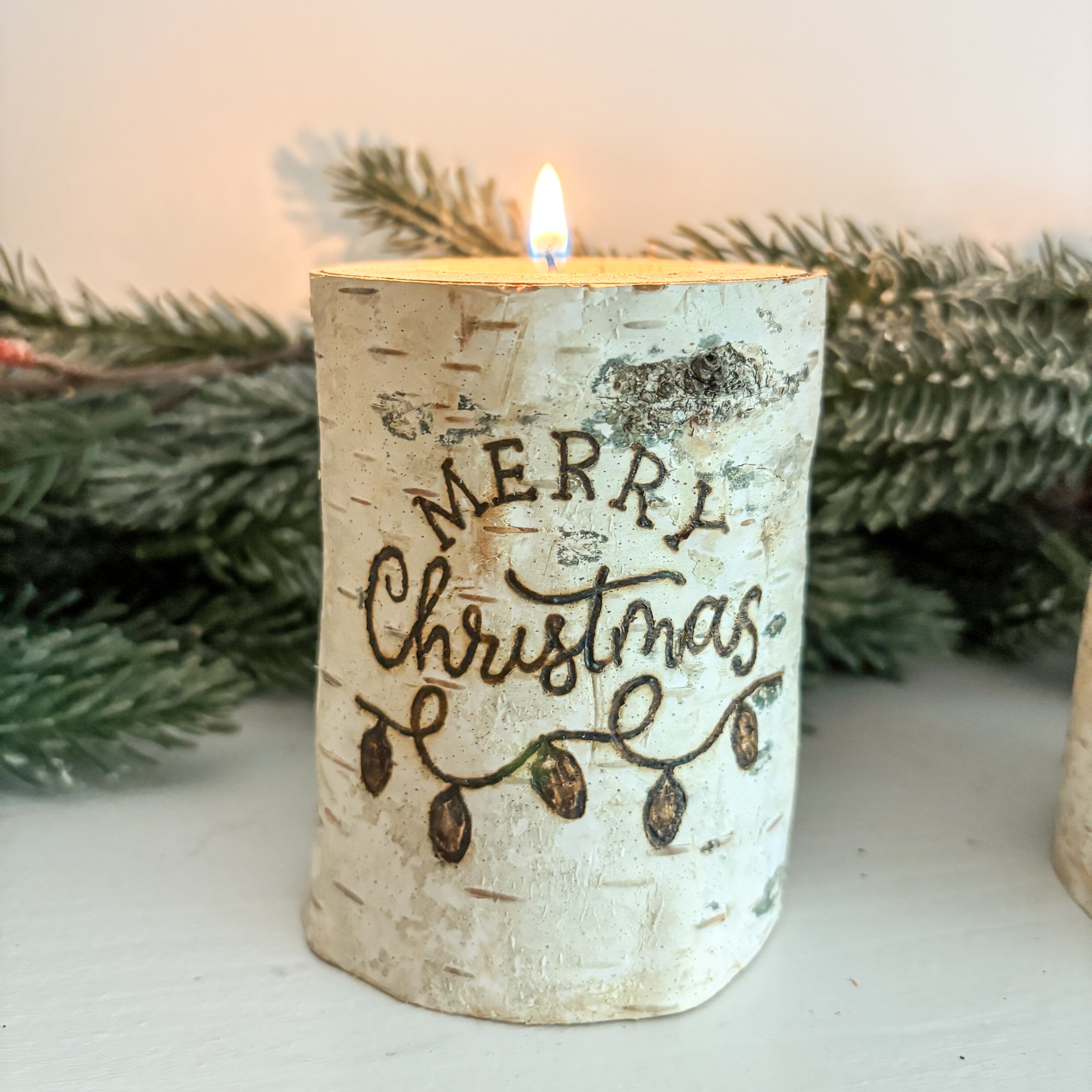 Candle with 'Merry Christmas' text on a white surface with greenery in the background