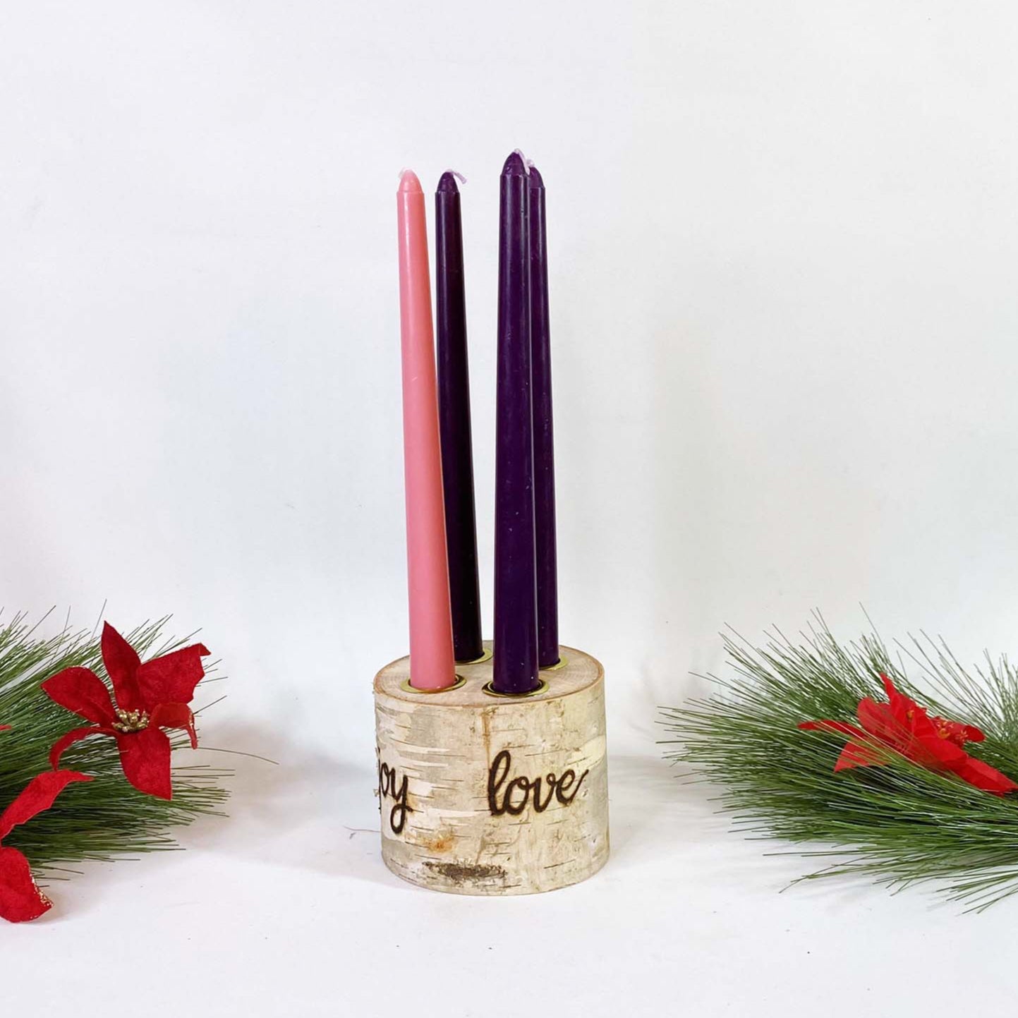 A birch wood advent wreath with four taper candles in purple and pink colors, placed on a white surface with red and green holiday decorations around it. The wreath has the words 'Hope', 'Peace', 'Joy', and 'Love' inscribed on it.