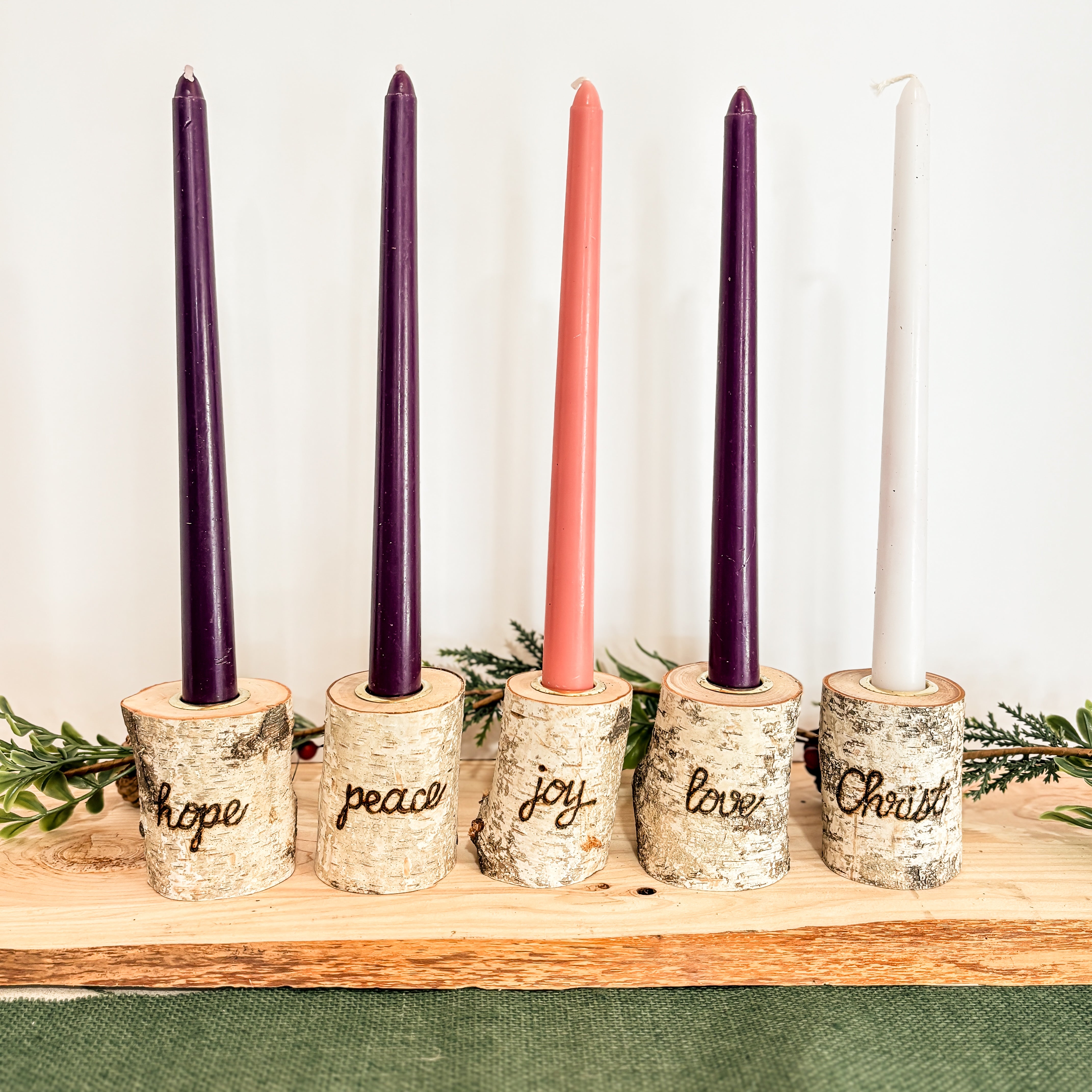 Set of five birch wood taper candle holders with engraved words, hope peace joy love Christ set up for Christmas with holiday greenery on a white background