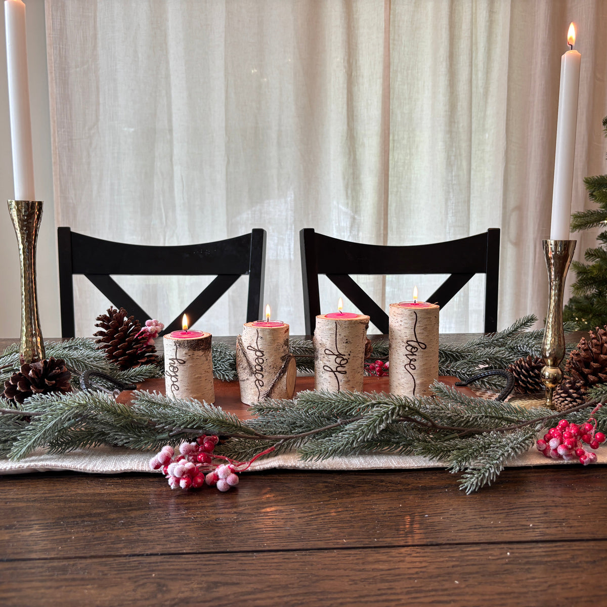 A festive holiday table centerpiece with four wrapped corks as candle holders, lit white taper candles in gold holders, pinecones, evergreen branches, and pink berries on a dark wood table with black chairs and a Christmas tree in the background.