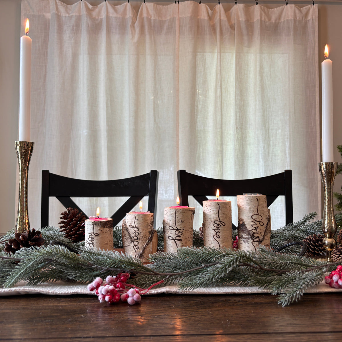A festive holiday table centerpiece with five lit taper candles wrapped in natural birch bark, surrounded by pine branches, pinecones, and pink berries on a dark wooden table. Two tall white taper candles in gold holders stand on either side, with black chairs and sheer curtains in the background.
