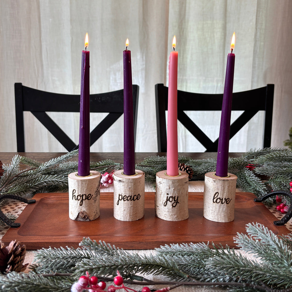 Four taper candles in purple and pink candle sleeves set in a row on a wooden tray, each mounted in a small cylindrical holder engraved with the words 'hope', 'peace', 'joy', and 'love'. The tray is decorated with pine branches, pinecones, and red berries on a wooden table, with black chairs and a cream curtain in the background.