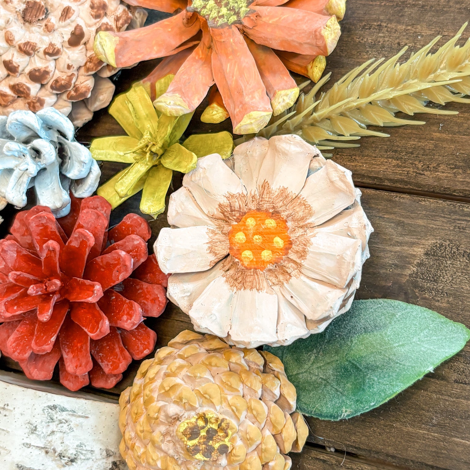 A collection of autumn-themed decorative craft items, including painted pinecones, a dried flower, a wheat stem, and leaves, arranged on a rustic wooden background.