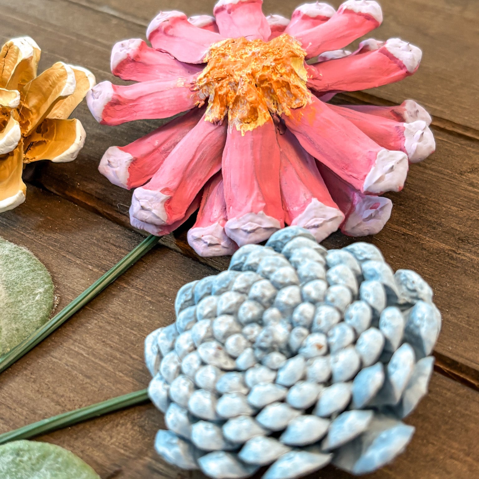 Hand-painted pinecone flowers displayed on a rustic wooden surface. The main flower in the center is painted pink with white-tipped petals and a textured orange-yellow center, while a second flower in the foreground is painted light blue.