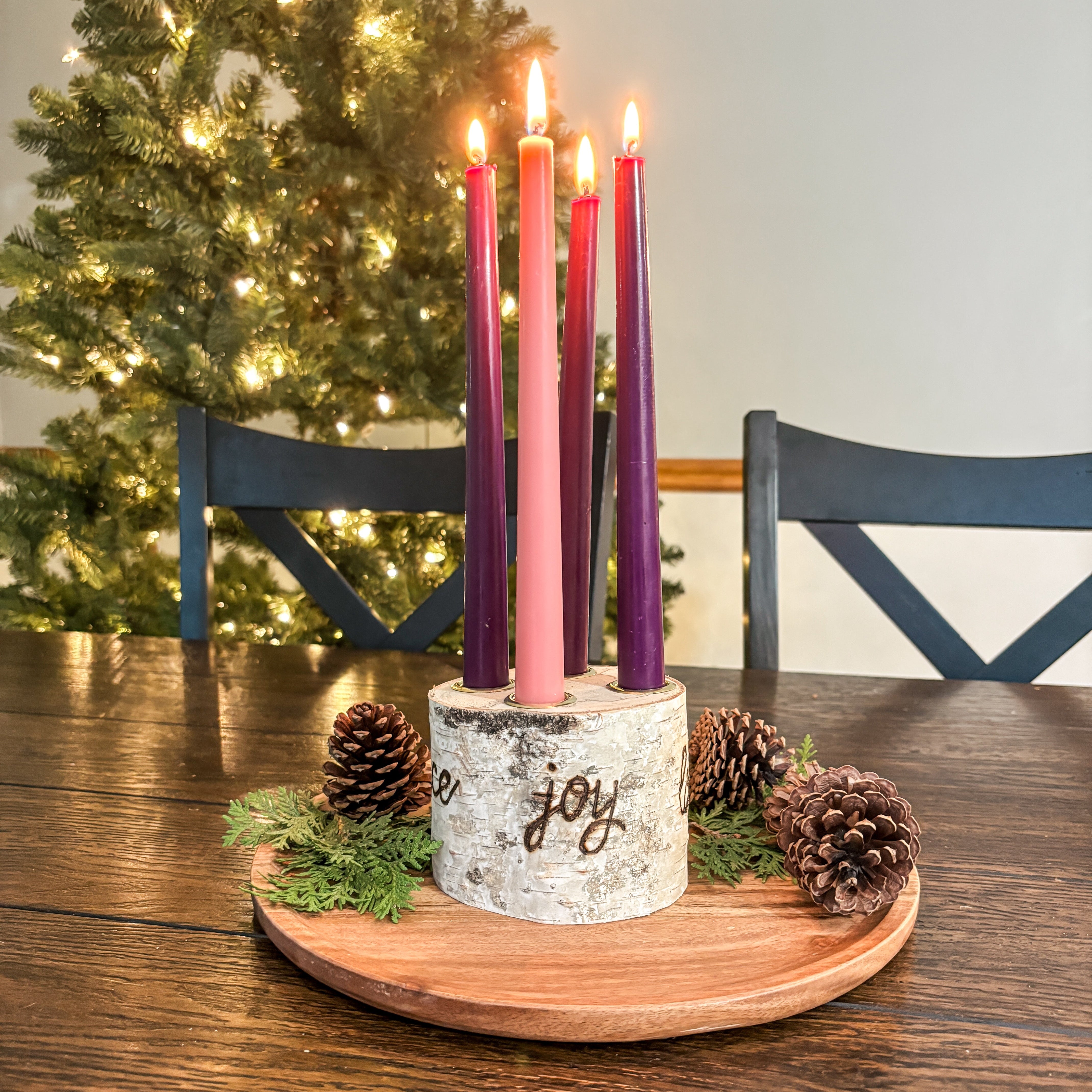 Decorative Advent wreath with lit candles on a wooden table in front of a Christmas tree.