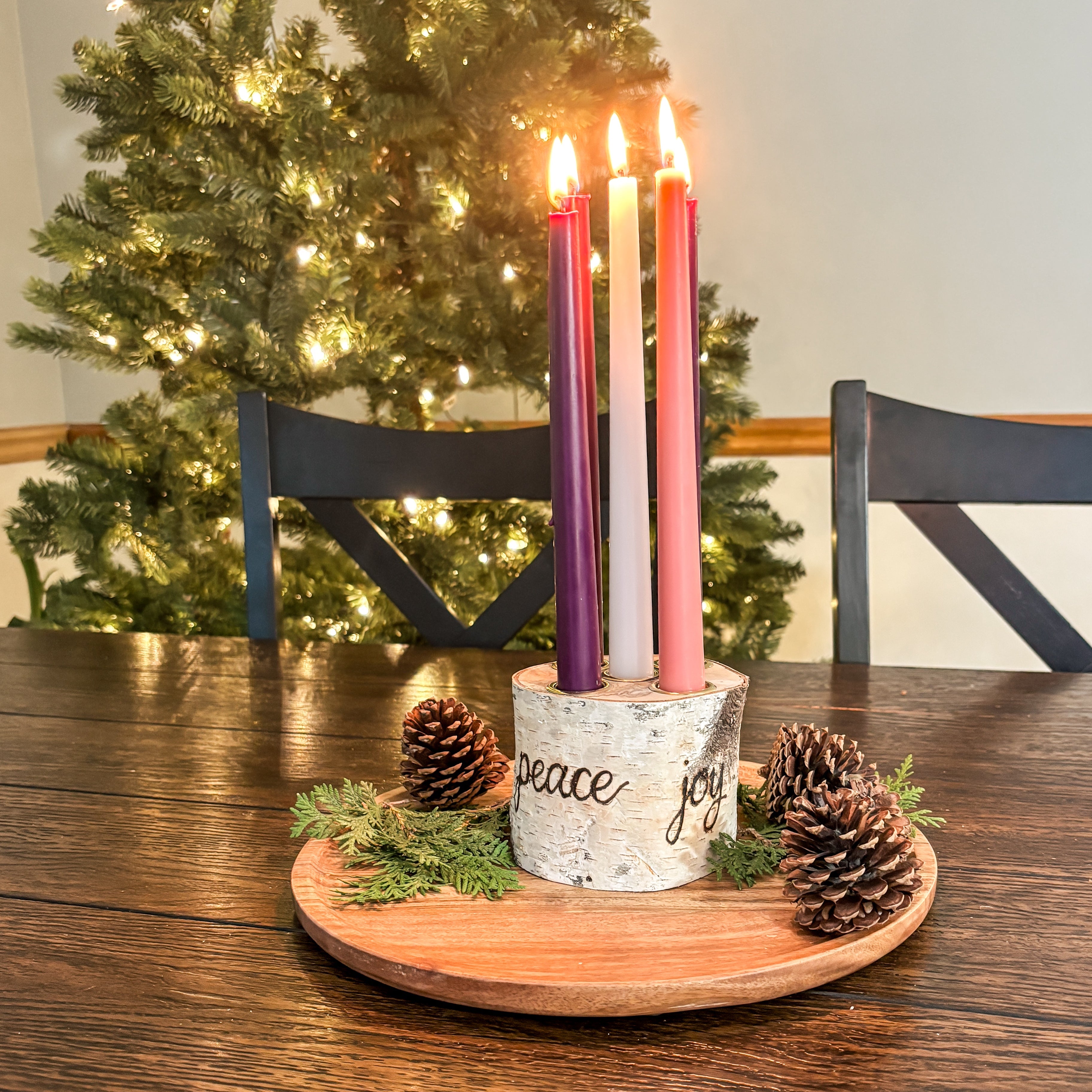 Decorative Advent wreath with lit candles on a wooden table in front of a Christmas tree.