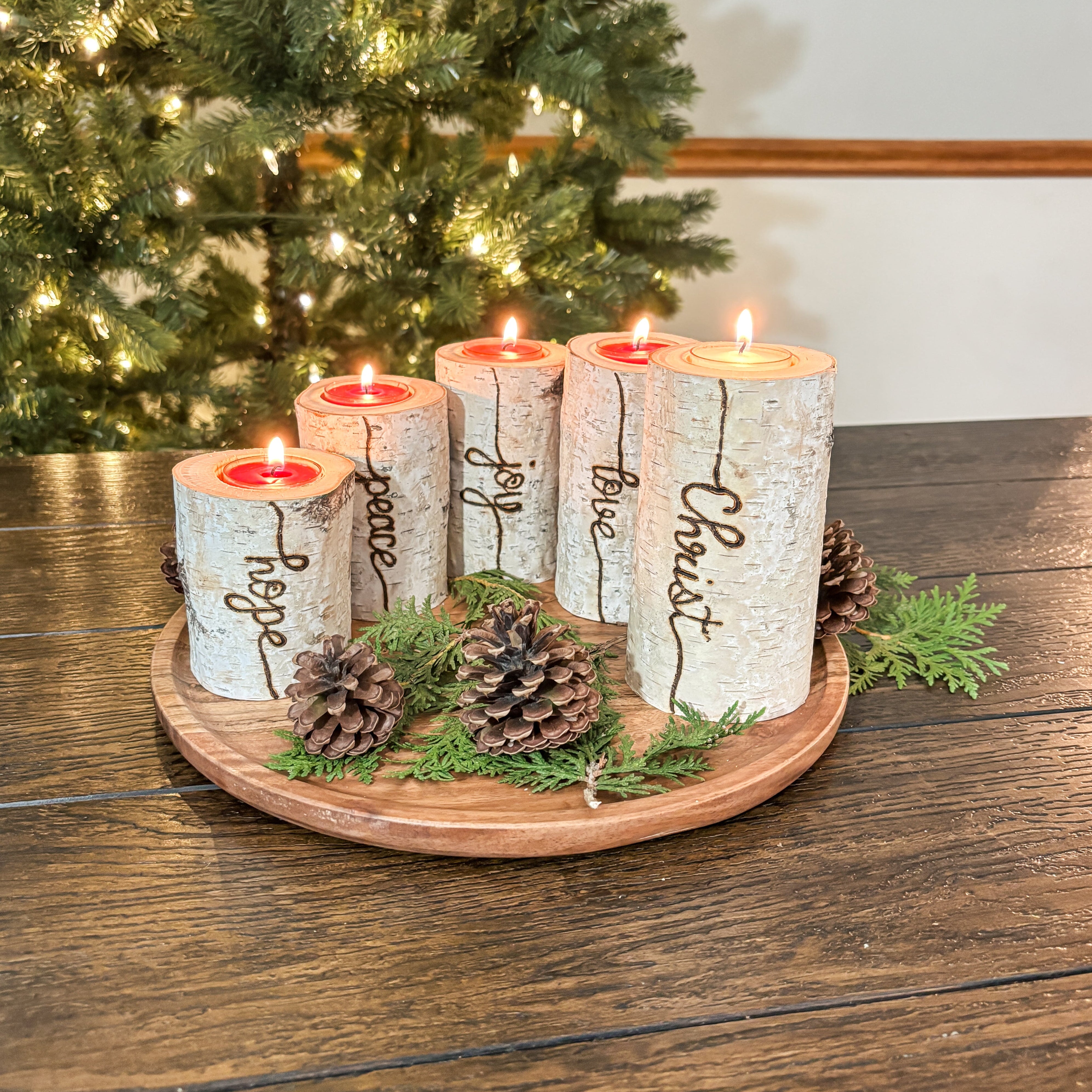 Decorative candles with engraved words on a wooden tray in front of a Christmas tree.