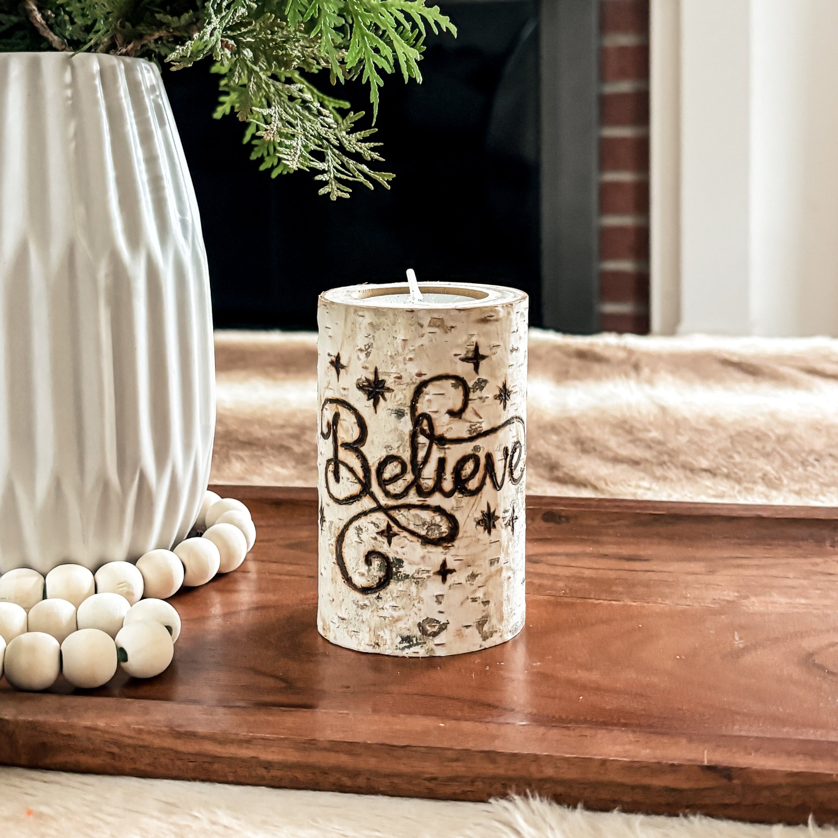 Decorative setup with birch candle hand burned with the word "Believe" and a vase of greenery, candle, and Christmas decorations in front of a fireplace.