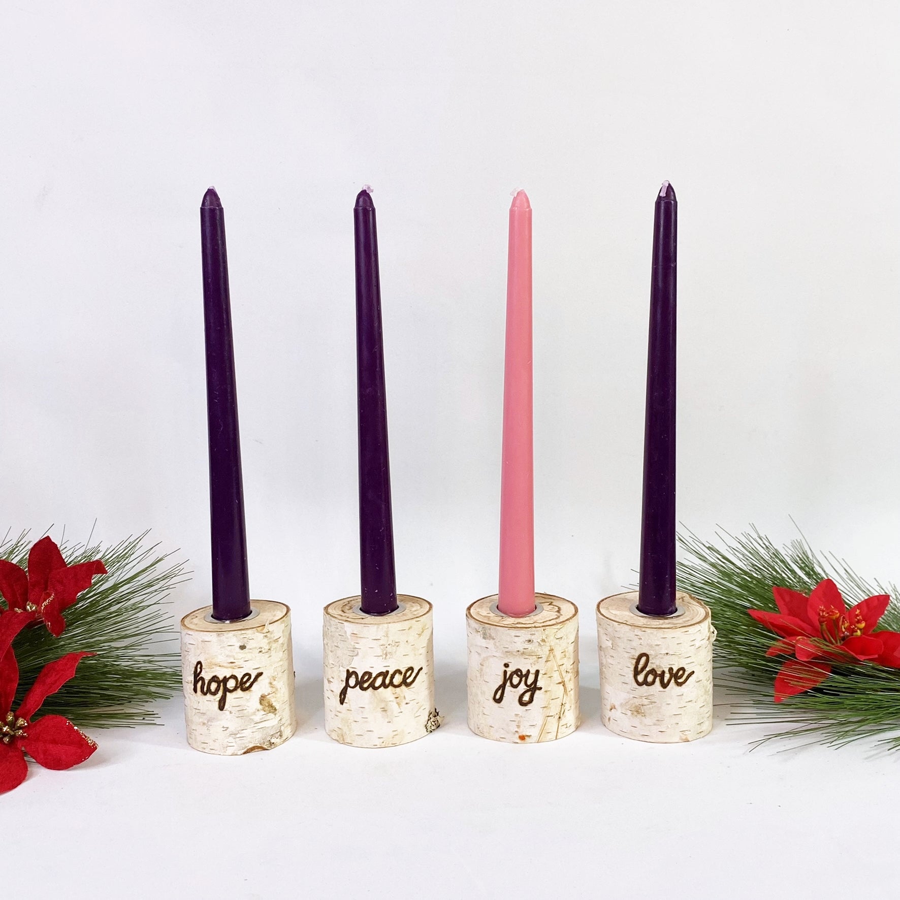 Four taper candles with birch wood holders, each labeled with a different word: Hope, Peace, Joy, Love. They are placed on a decorative wreath with red flowers and green pine needles.