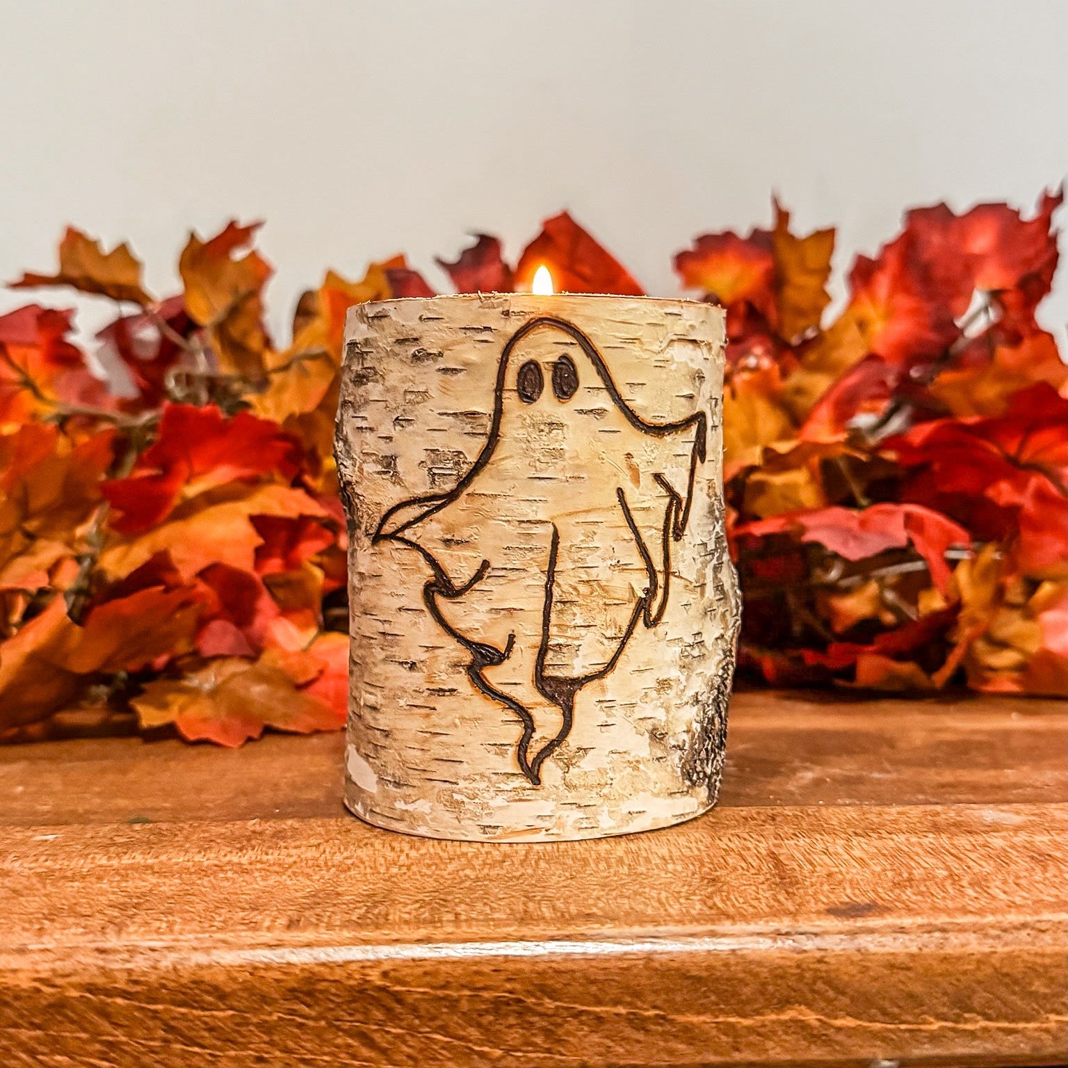 Birch bark candle holder with ghost design on a wooden surface with autumn leaves in the background