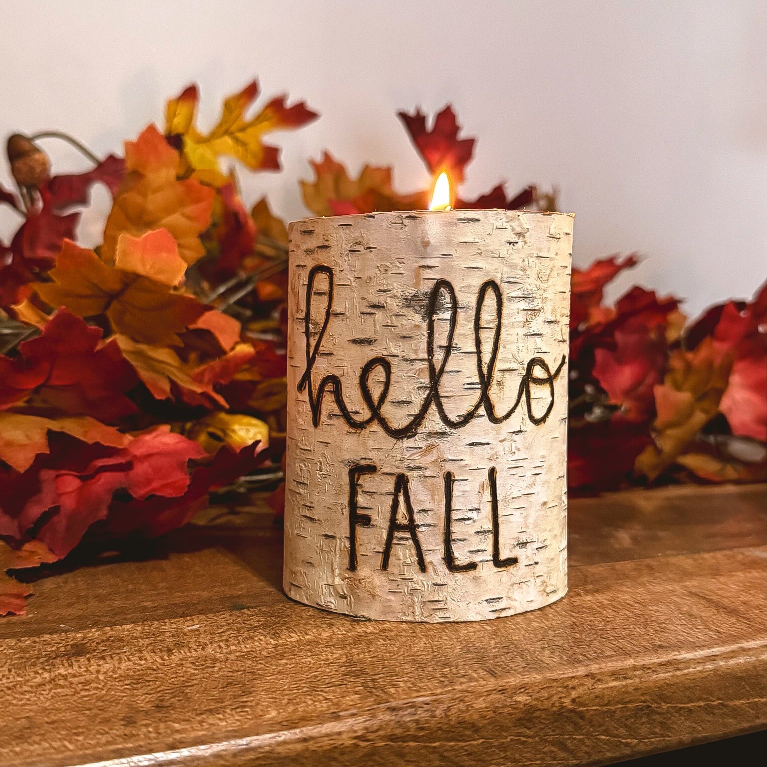 Candle with 'hello FALL' text on a wooden surface with autumn leaves.