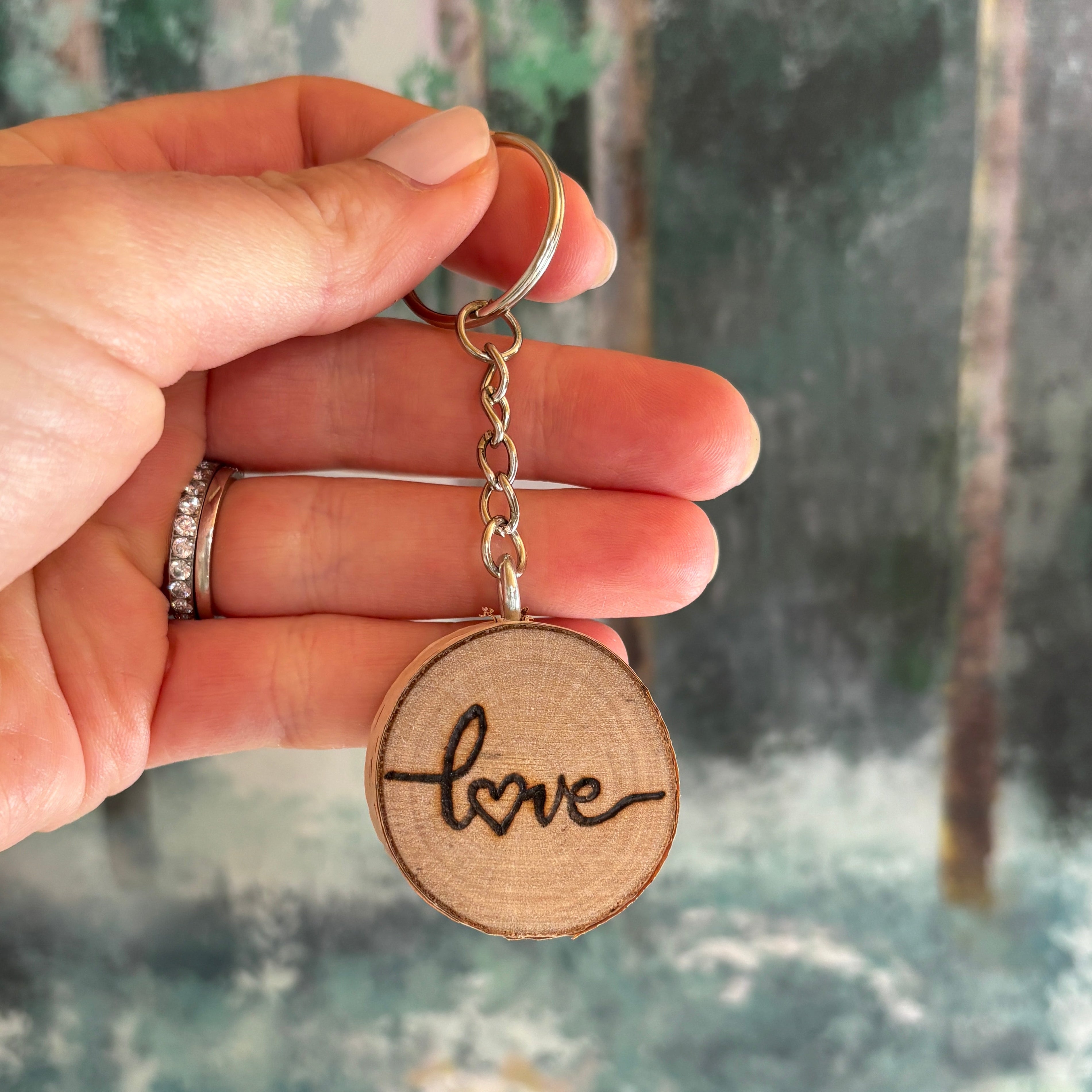 Hand holding a wooden round charm with 'love' engraved, chain, and ring against a blurred background