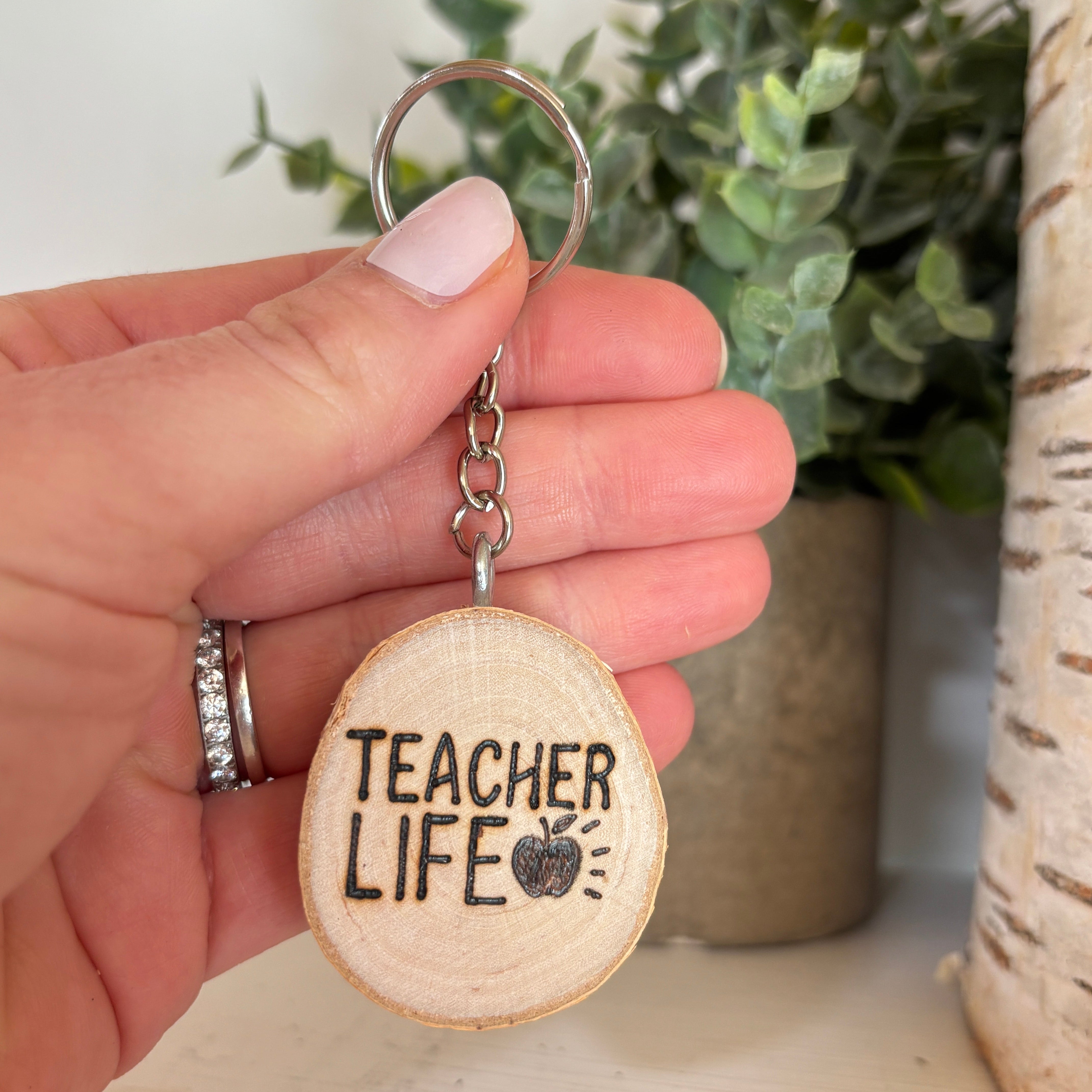 Hand holding a keychain with 'Teacher Life' text and an apple icon, with a plant and vase in the background.