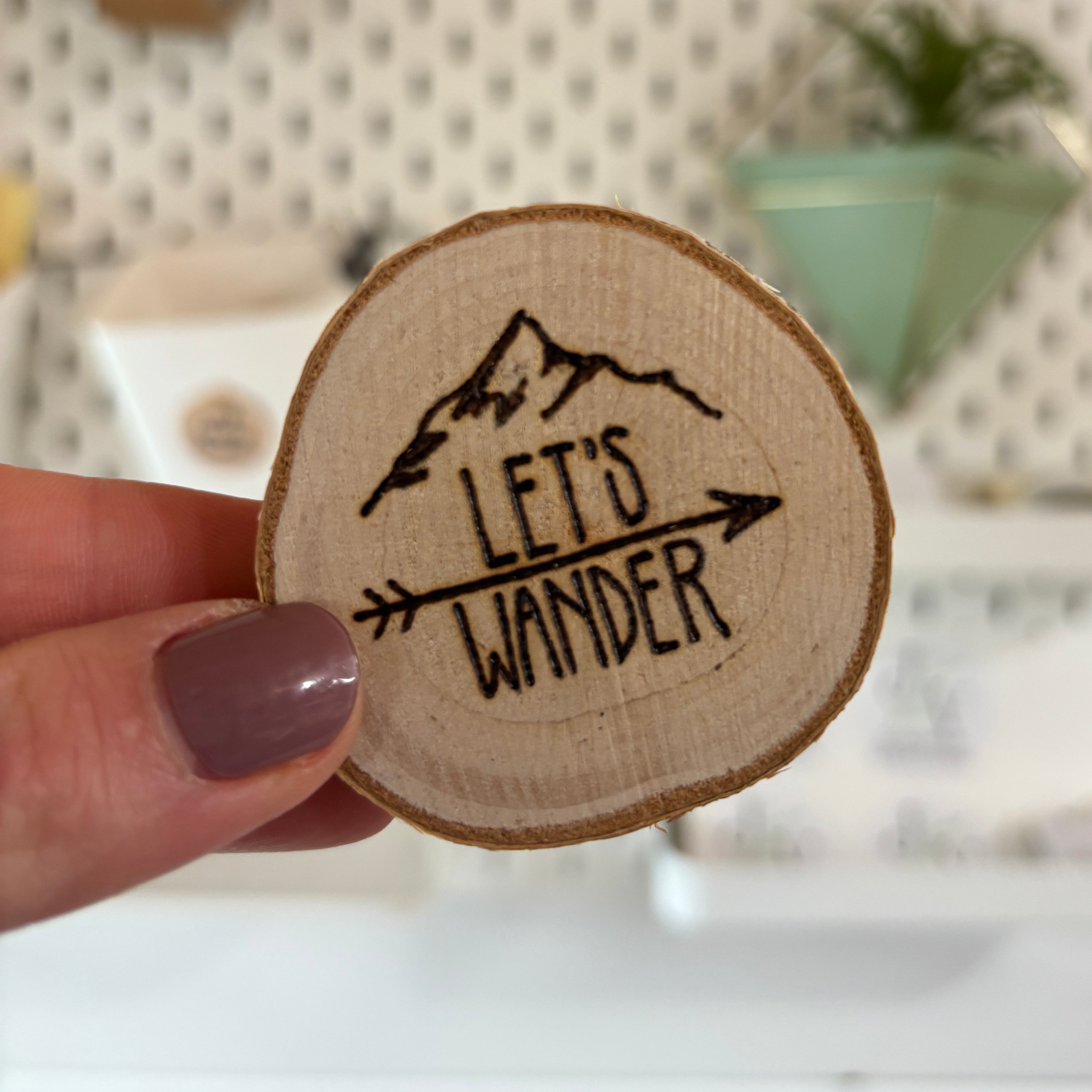 A hand with mauve nail polish holds a round, natural wood slice magnet featuring a black engraved mountain and the words 'LET'S WANDER' with an arrow. The background is a brightly lit, softly blurred craft room or studio space with a pegboard.