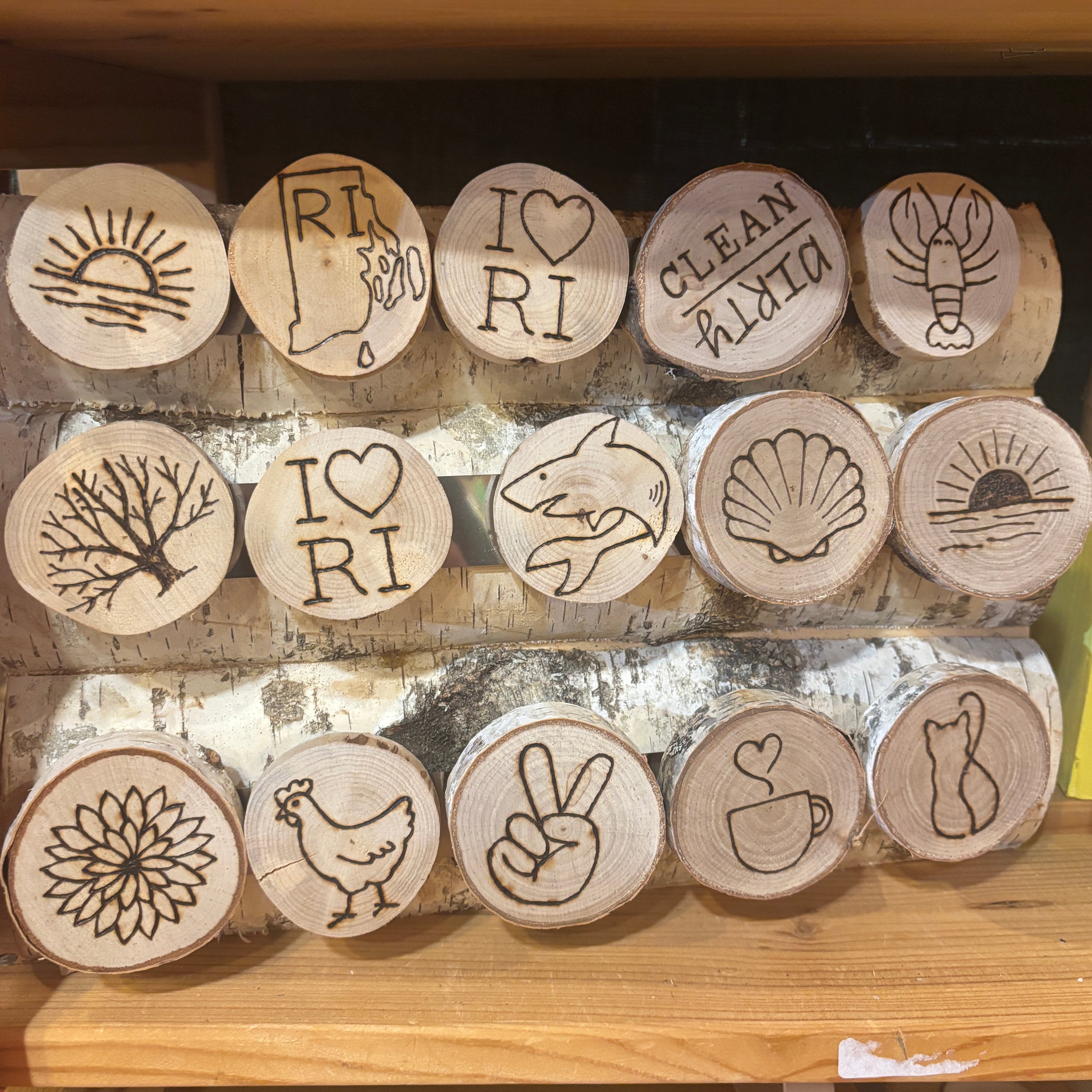 A set of round wooden coasters with engraved beach and nature-themed designs, including a lobster, seashell, shark, dahlia, chicken, peace sign, coffee cup, and map of Rhode Island, displayed on a wooden shelf.
