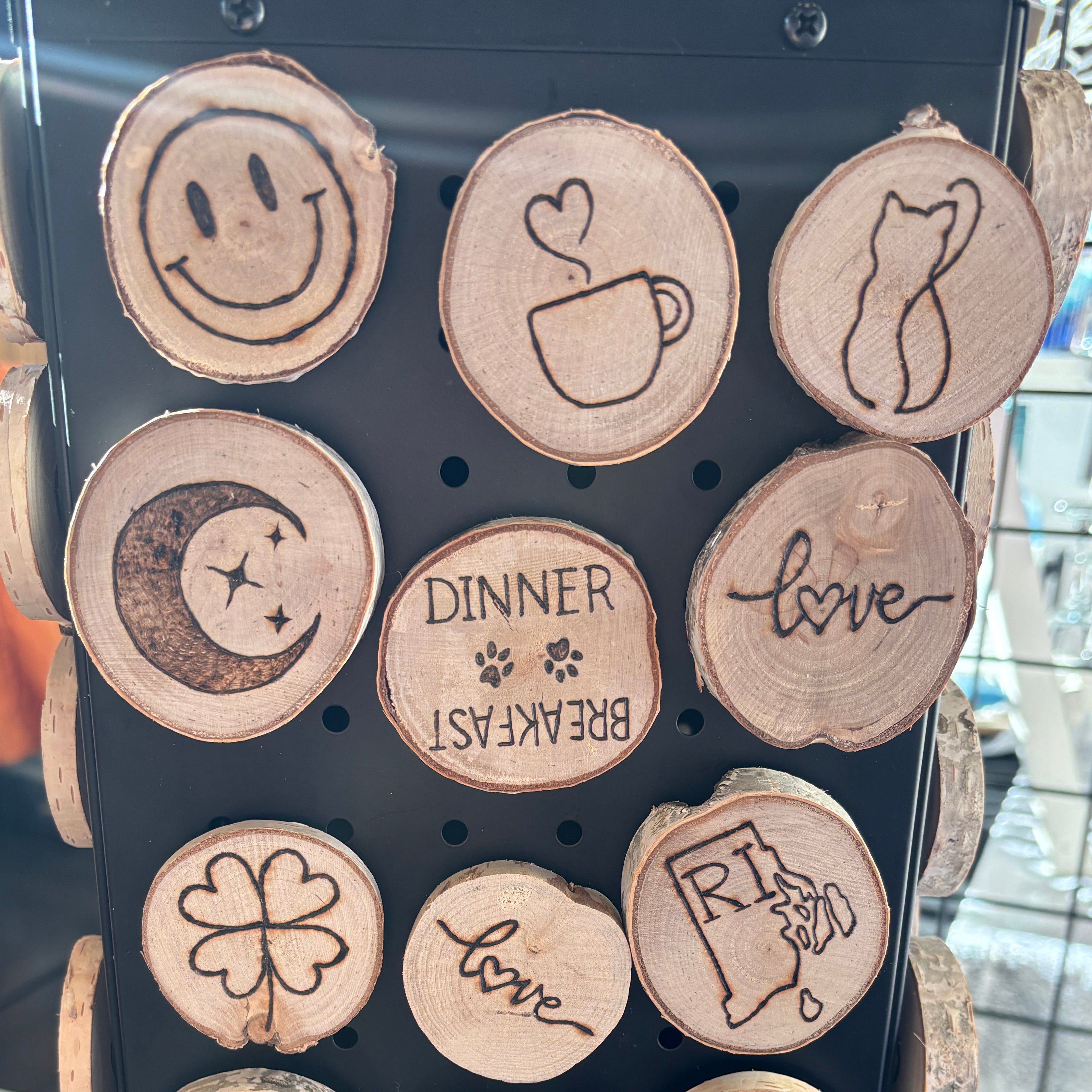 A black pegboard wall sign decorated with multiple round natural wood slice magnets, each engraved or burned with a different decorative symbol or phrase such as smiley face, coffee cup with heart, cat silhouette, moon, dinner breakfast, love, clover, RI flag, dirty clean, joy, my kitchen my rules, boot, peace sign, and crescent moon.