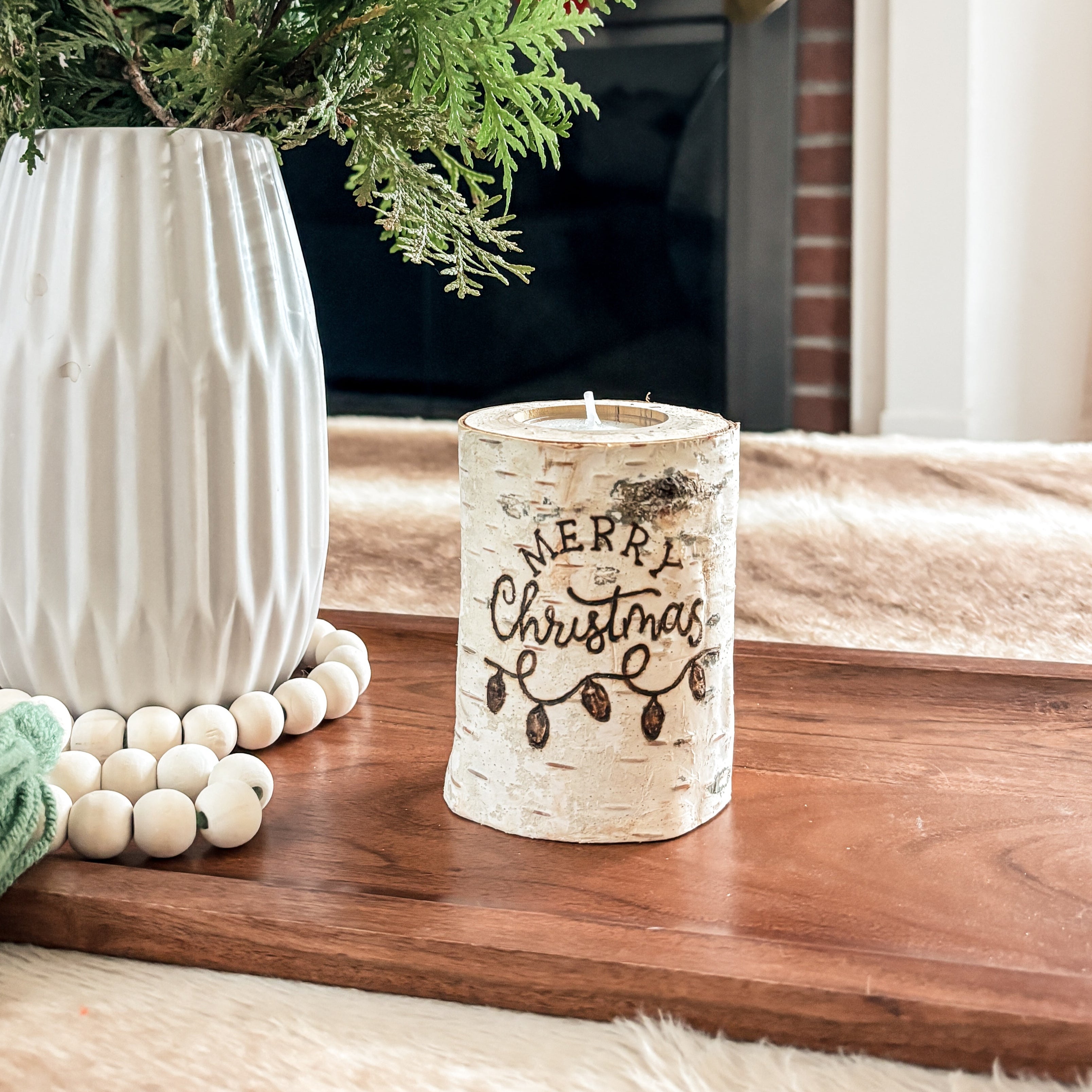 Decorative setup with a vase of greenery, candle, and Christmas stockings in the background and a Christmas birch wood candle as decoration on a tray 