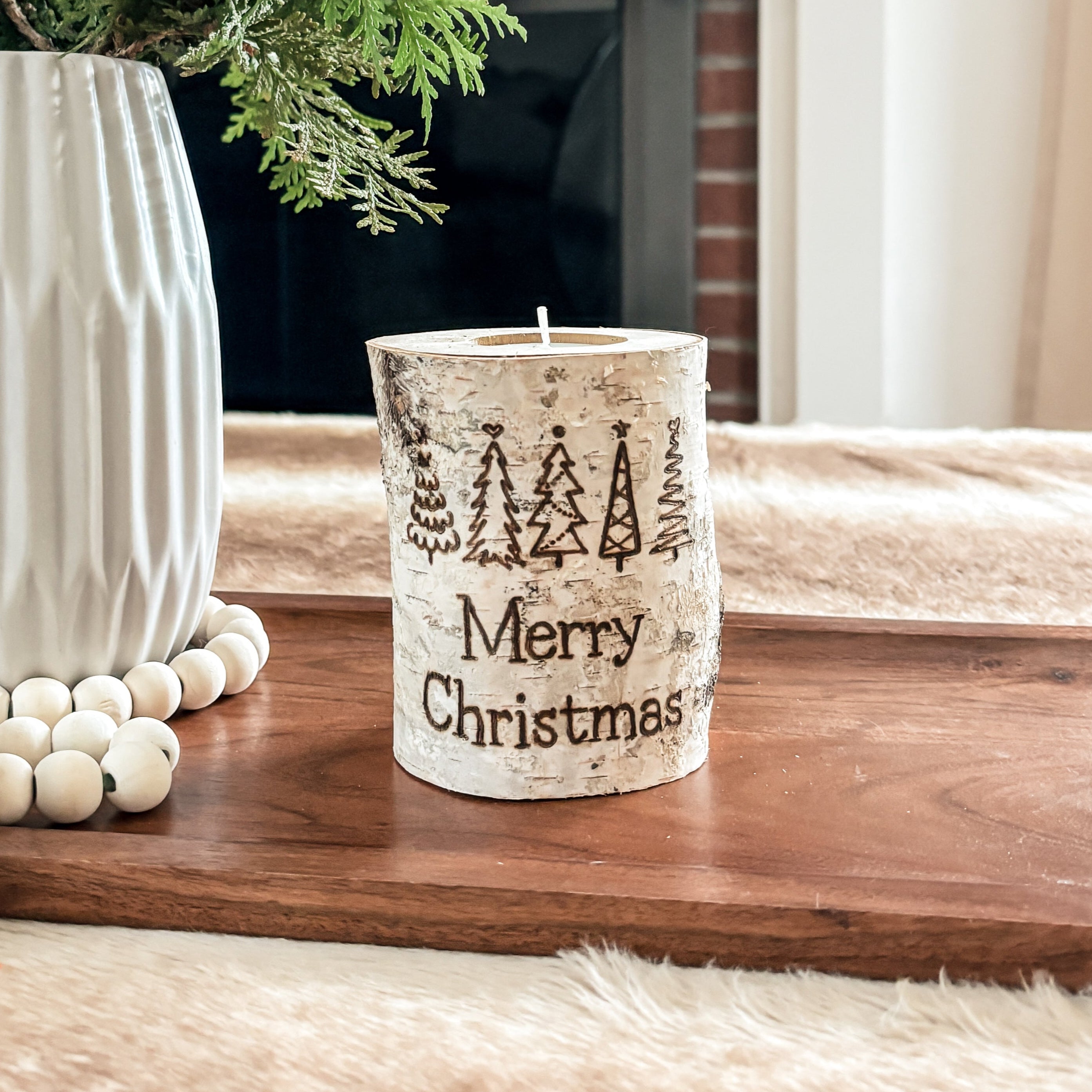 Decorative Christmas scene with a birch wood candle that says Merry Christmas, a vase, candle, and greenery on a mantelpiece.