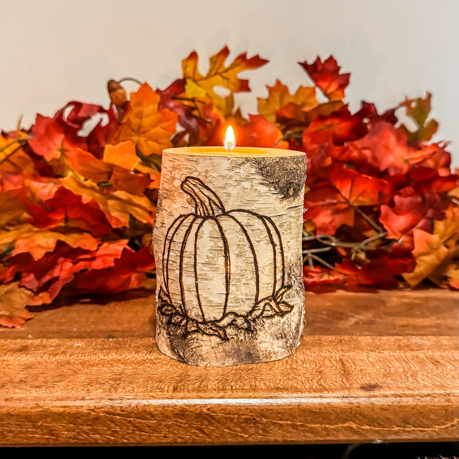 Candle with pumpkin design in front of autumn leaves on a wooden surface