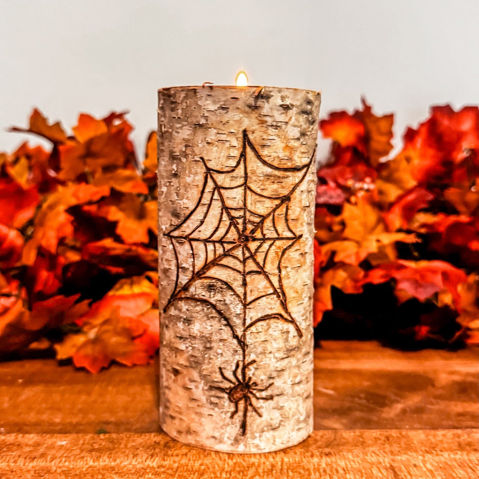 Decorative candle with spider web design on a wooden surface with autumn leaves in the background