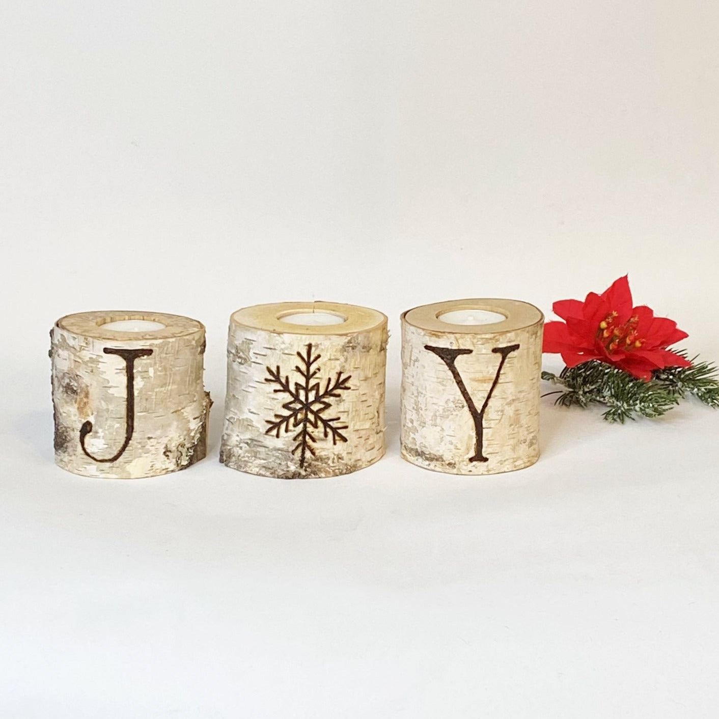 Three rustic birch wood candle holders with the word 'JOY' burned on them, placed on a white background with a red poinsettia flower to the side.